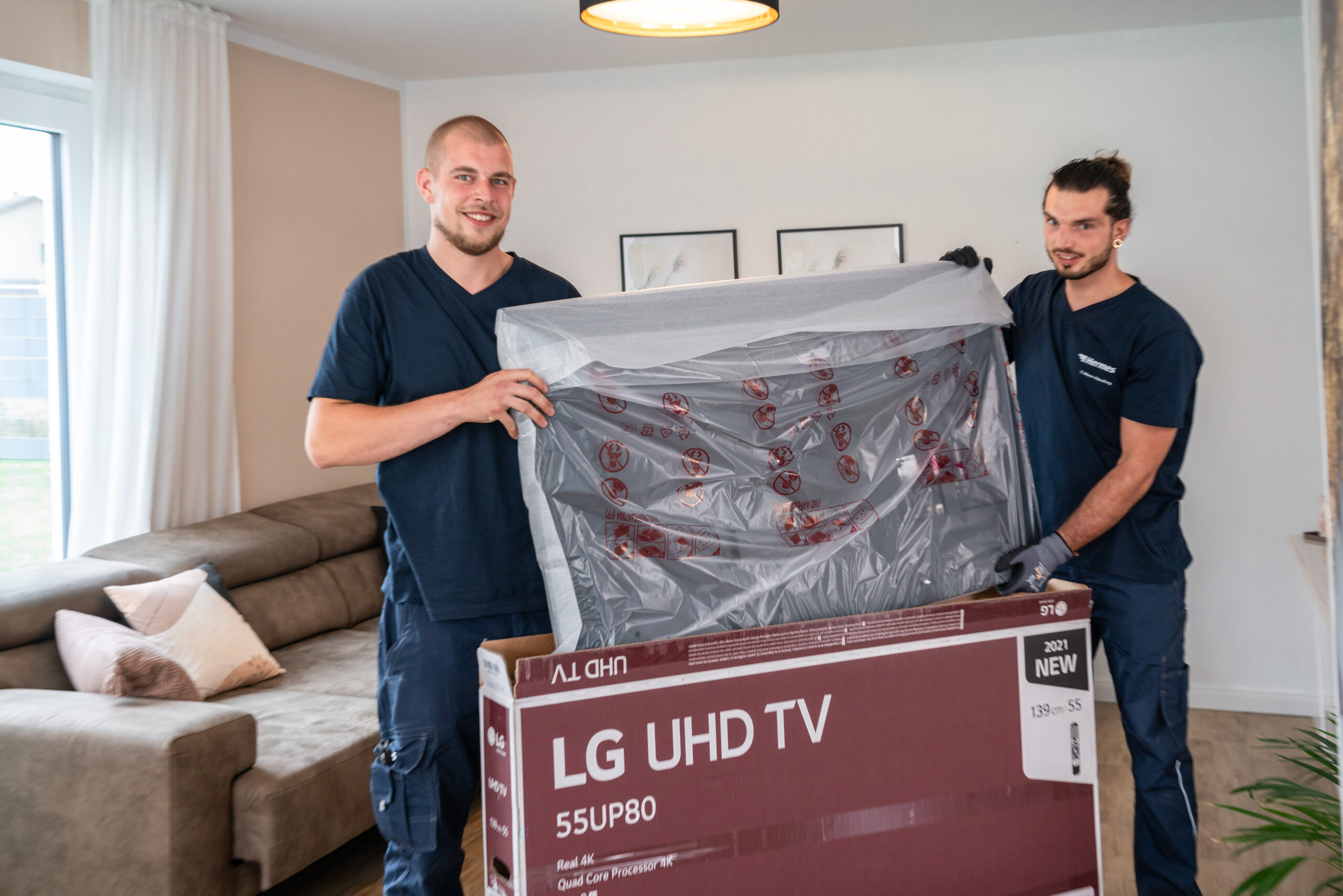 Zwei HES-Mitarbeiter tragen einen verpackten Fernseher in eine Wohnung.
